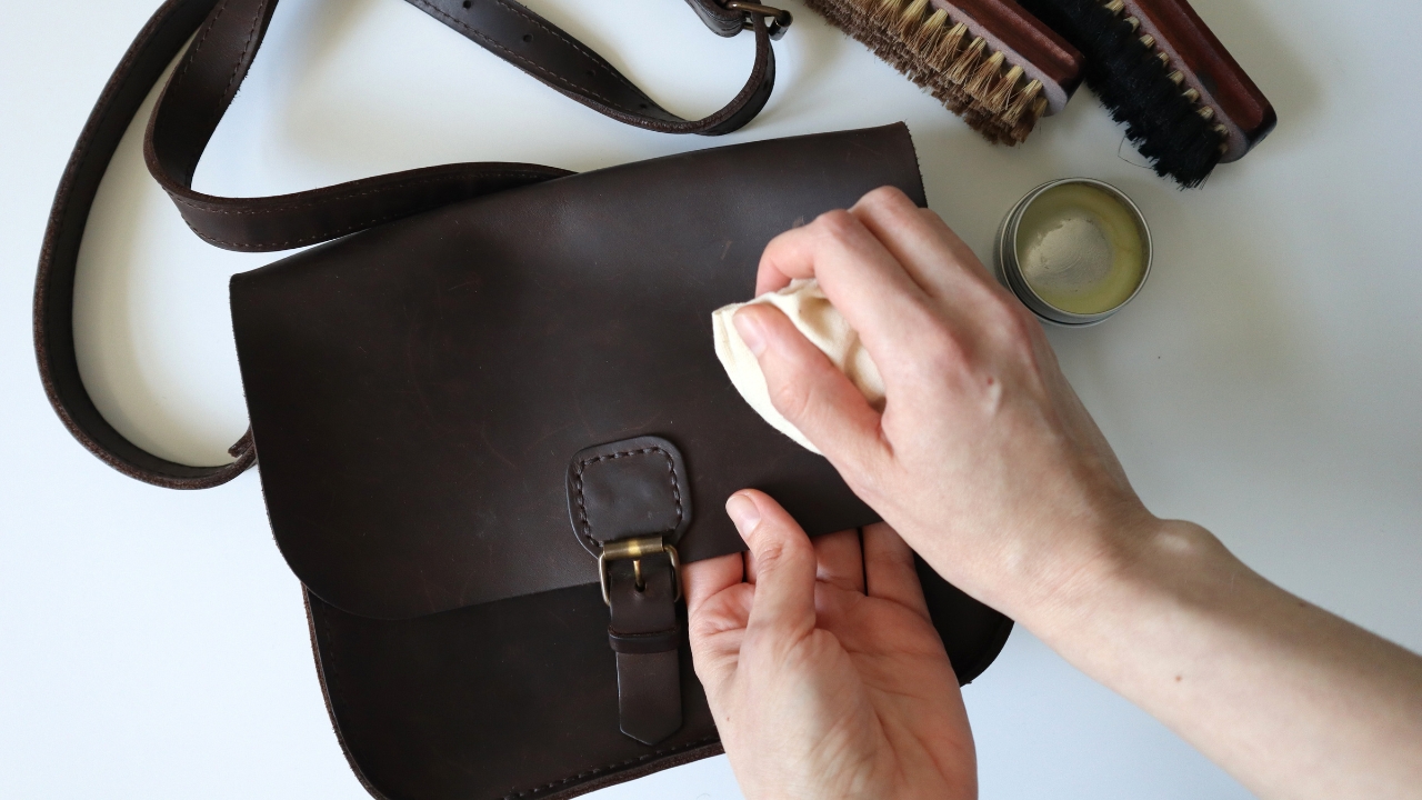 Person wipes a stain off a dark leather bag with a cloth next to care tools on a table